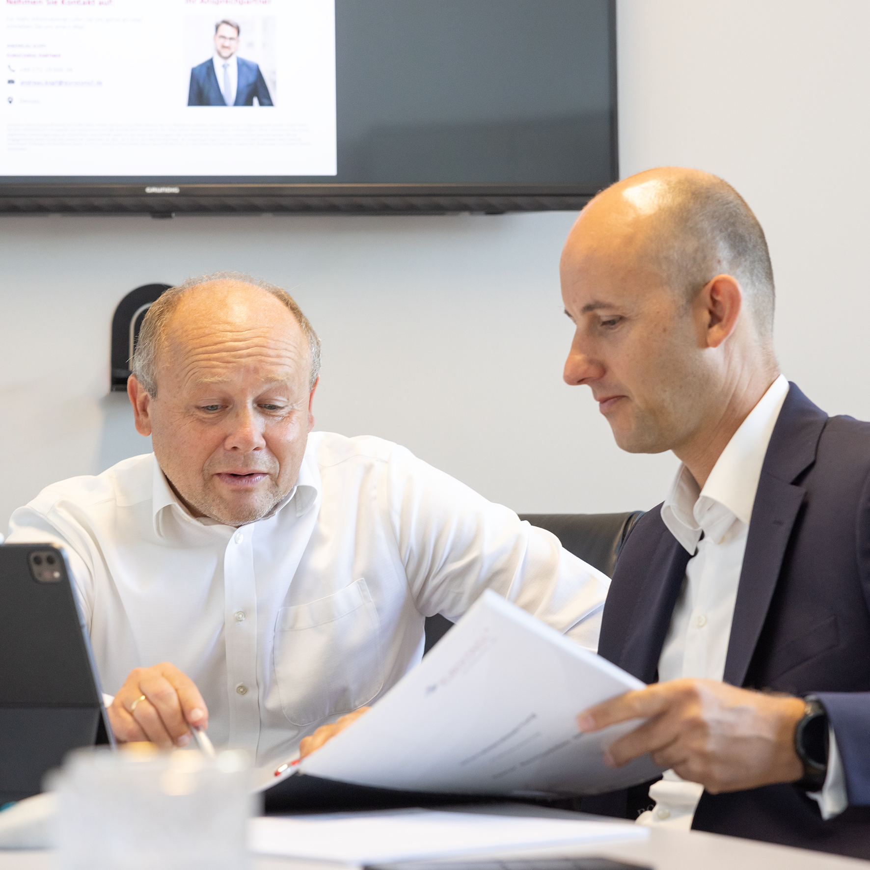 Zwei Männer besprechen Dokumente in einem Büro, einer hält einen Stift und schaut auf ein Tablet.
