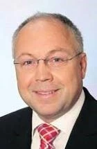 Ein Mann mit Brille, dunklem Anzug und kariertem Krawatten-Tuch lächelt.